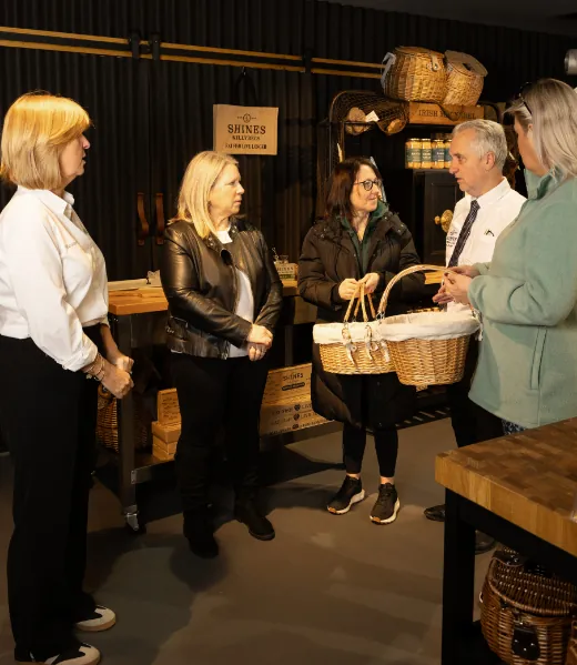 Five adults stand in a shop with wicker baskets and products on shelves. Two women hold a large wicker basket together whilst talking to a man in a white shirt. The atmosphere is friendly and casual.