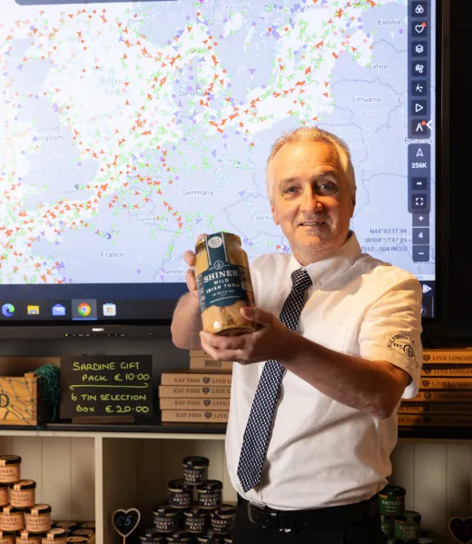 A man in a white shirt and tie holds a jar of Shine’s seafood in front of a large screen displaying a map with coloured markers. Shelves with jars and price signs are visible beside him.