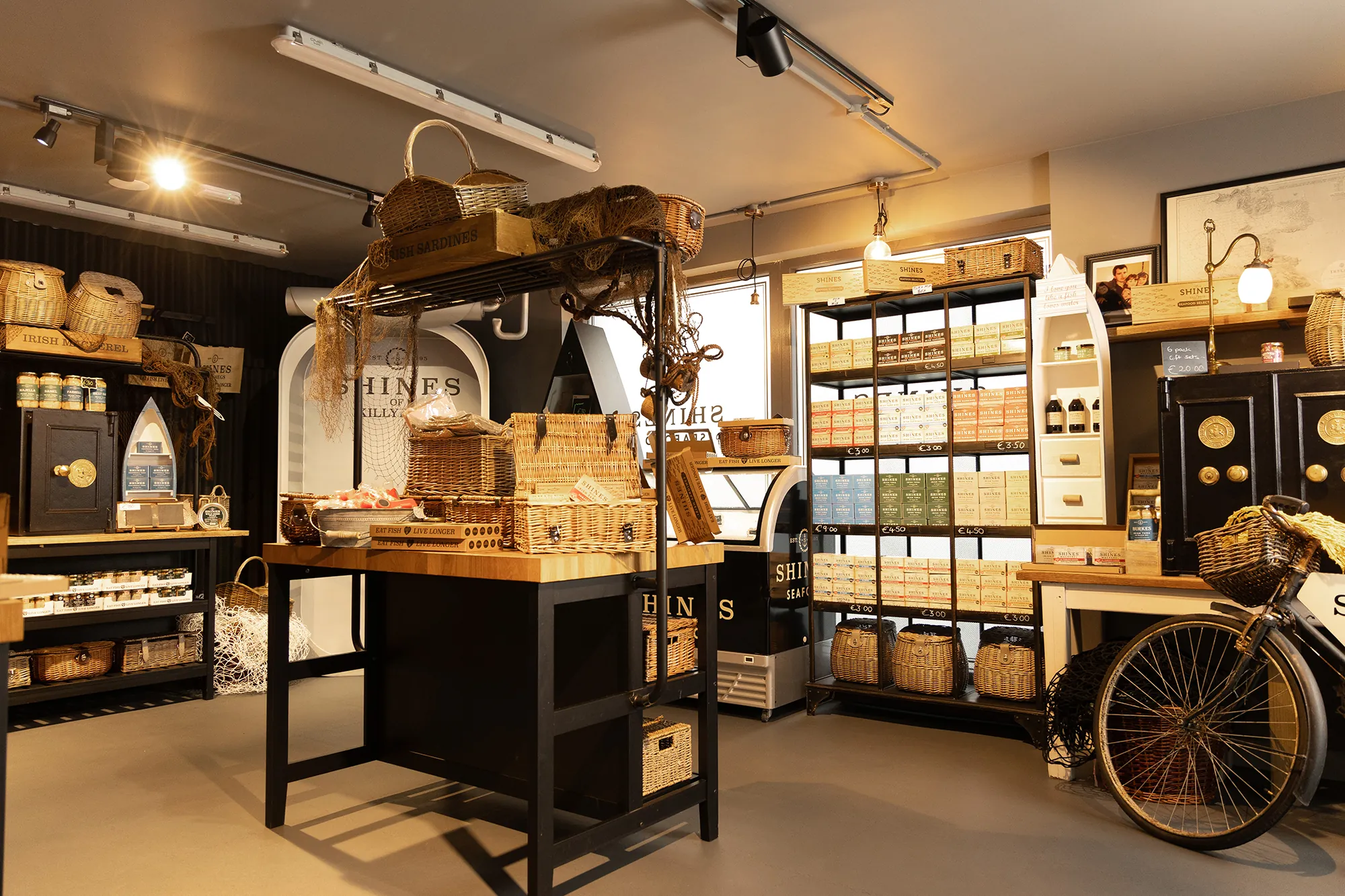 A cosy shop interior with shelves and tables displaying woven baskets, boxes, and gourmet food items. Warm lighting highlights rustic décor, a vintage bicycle, and neatly organised goods on dark wooden furniture.