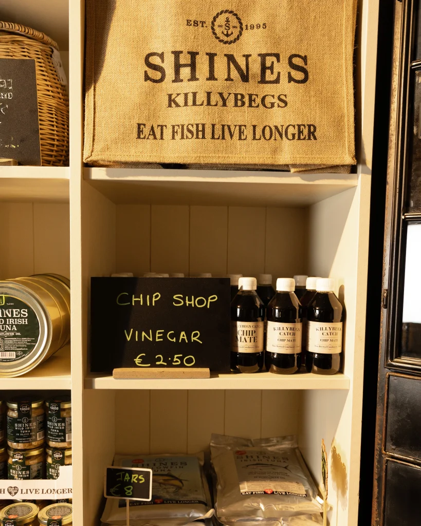 A shelf display featuring bottles of chip shop vinegar priced at €2.50, tins of seafood products, and a hessian bag with the text "SHINES Killybegs EAT FISH LIVE LONGER.