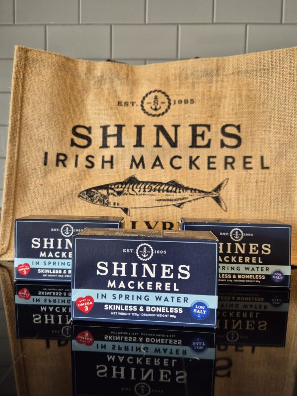Three boxes of Shines Mackerel Fillets in Spring Water - Low Salt 125g are shown in front of a hessian bag with "SHINES IRISH MACKEREL" and a fish illustration, set against a white tiled background.