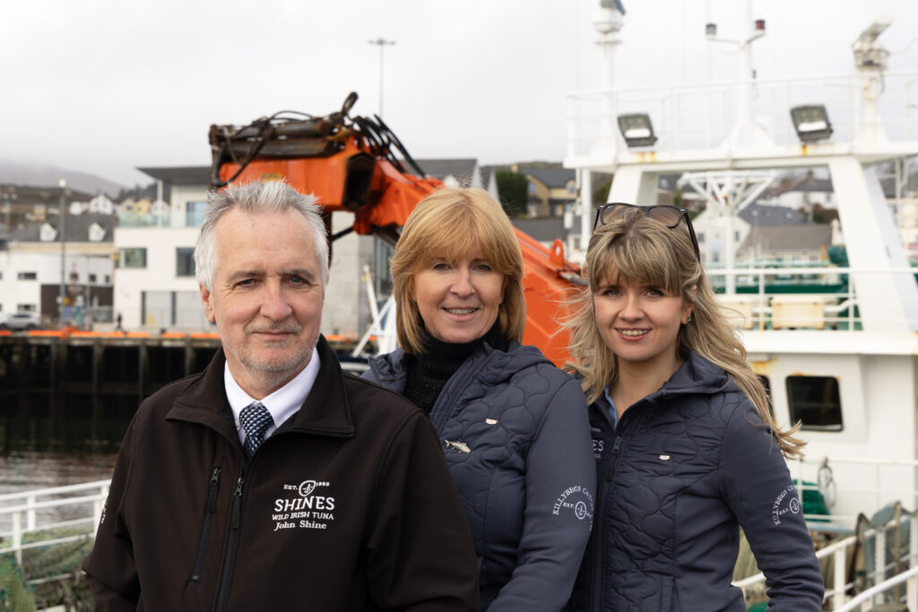 Three people wearing branded jackets stand on a quay in front of a fishing boat with an orange crane. Buildings and water are visible in the background. They are smiling and facing the camera.