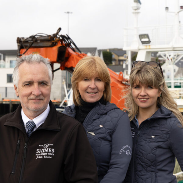 Three people wearing branded jackets stand on a quay in front of a fishing boat with an orange crane. Buildings and water are visible in the background. They are smiling and facing the camera.