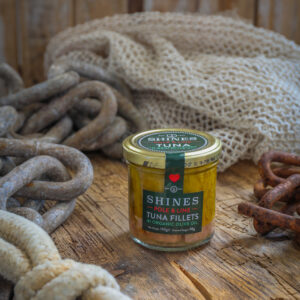 A glass jar of Shines Skipjack Tuna - Line and Pole - ORGANIC extra virgin olive oil rests on a rustic wooden surface, surrounded by thick rope, metal chains, and a piece of fishing net.