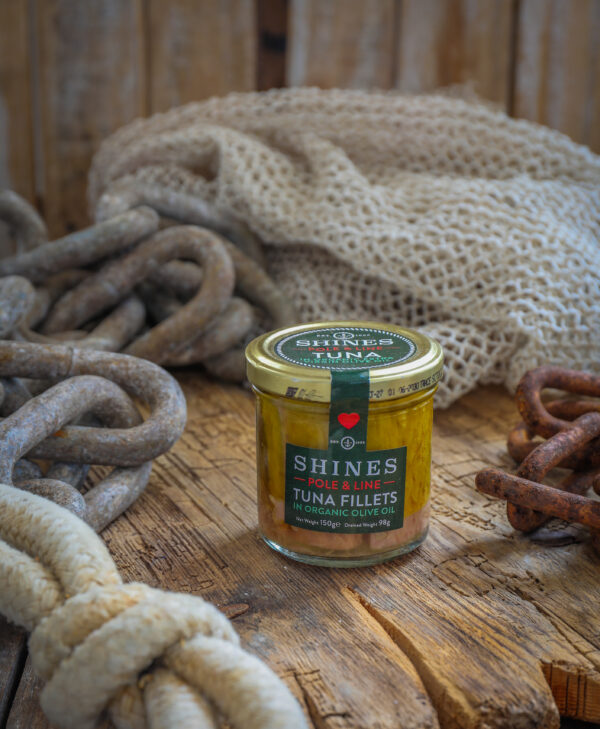 A glass jar of Shines Skipjack Tuna - Line and Pole - ORGANIC extra virgin olive oil rests on a rustic wooden surface, surrounded by thick rope, metal chains, and a piece of fishing net.