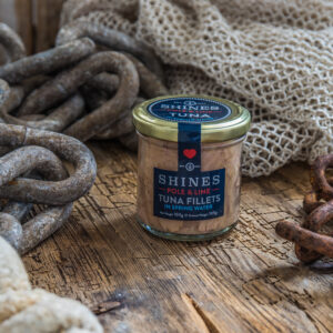 A glass jar of Shines Skipjack Tuna - Line and Pole in ORGANIC extra virgin olive oil rests on a rustic wooden surface, surrounded by coiled ropes, fishing nets, and rusty chains for a nautical feel.