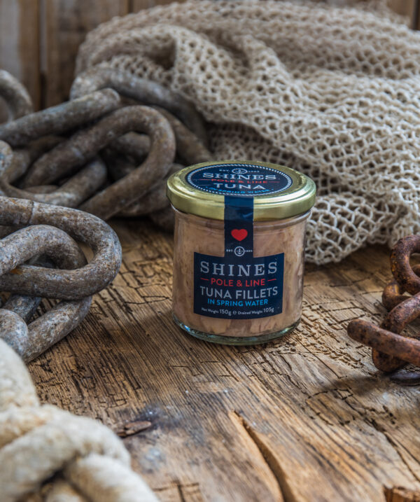 A glass jar of Shines Skipjack Tuna - Line and Pole in ORGANIC extra virgin olive oil rests on a rustic wooden surface, surrounded by coiled ropes, fishing nets, and rusty chains for a nautical feel.