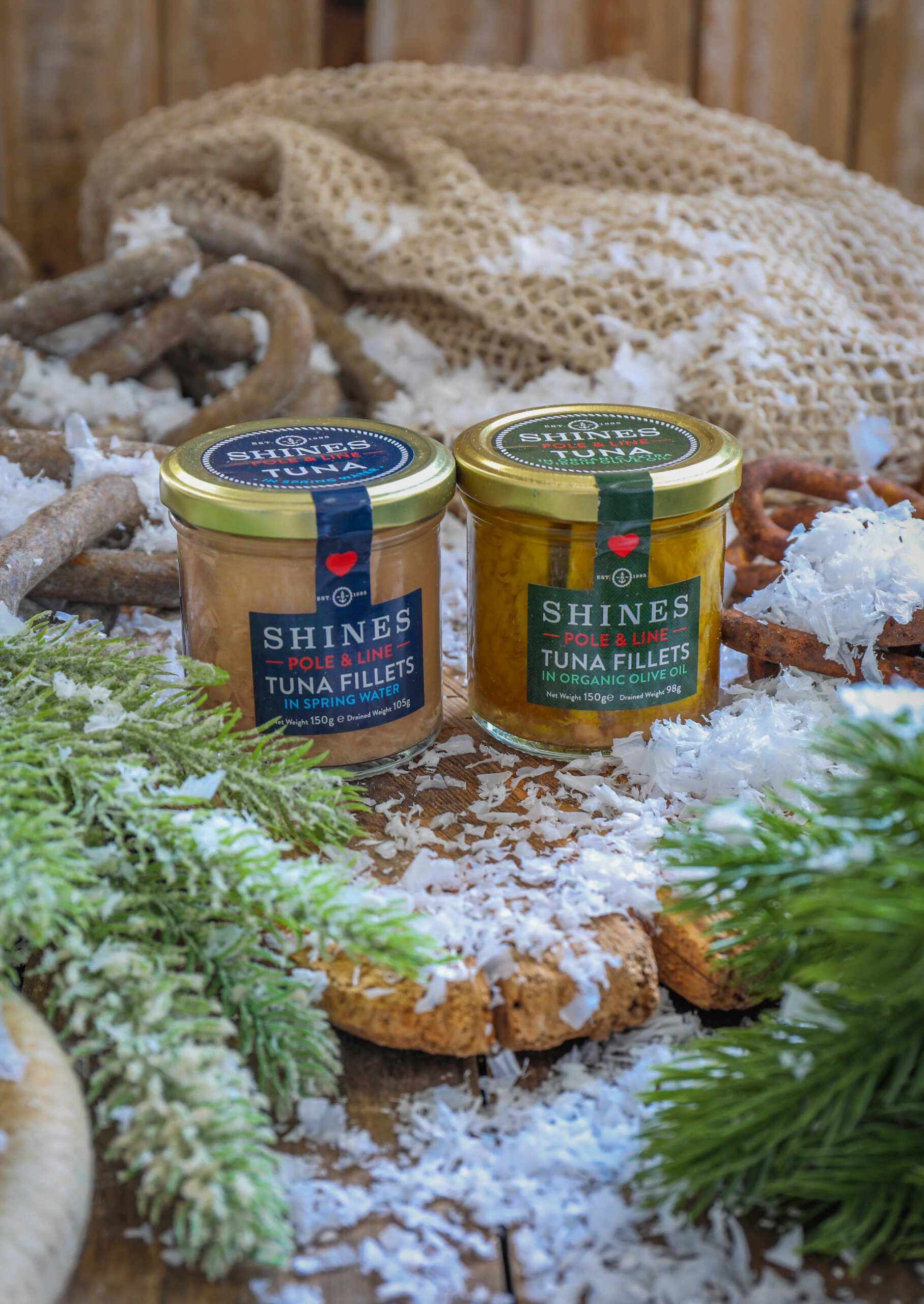 Two jars of Shines tuna fillets sit on a rustic surface surrounded by artificial snow, greenery, a fishing net, and rope, creating a festive and nautical atmosphere.