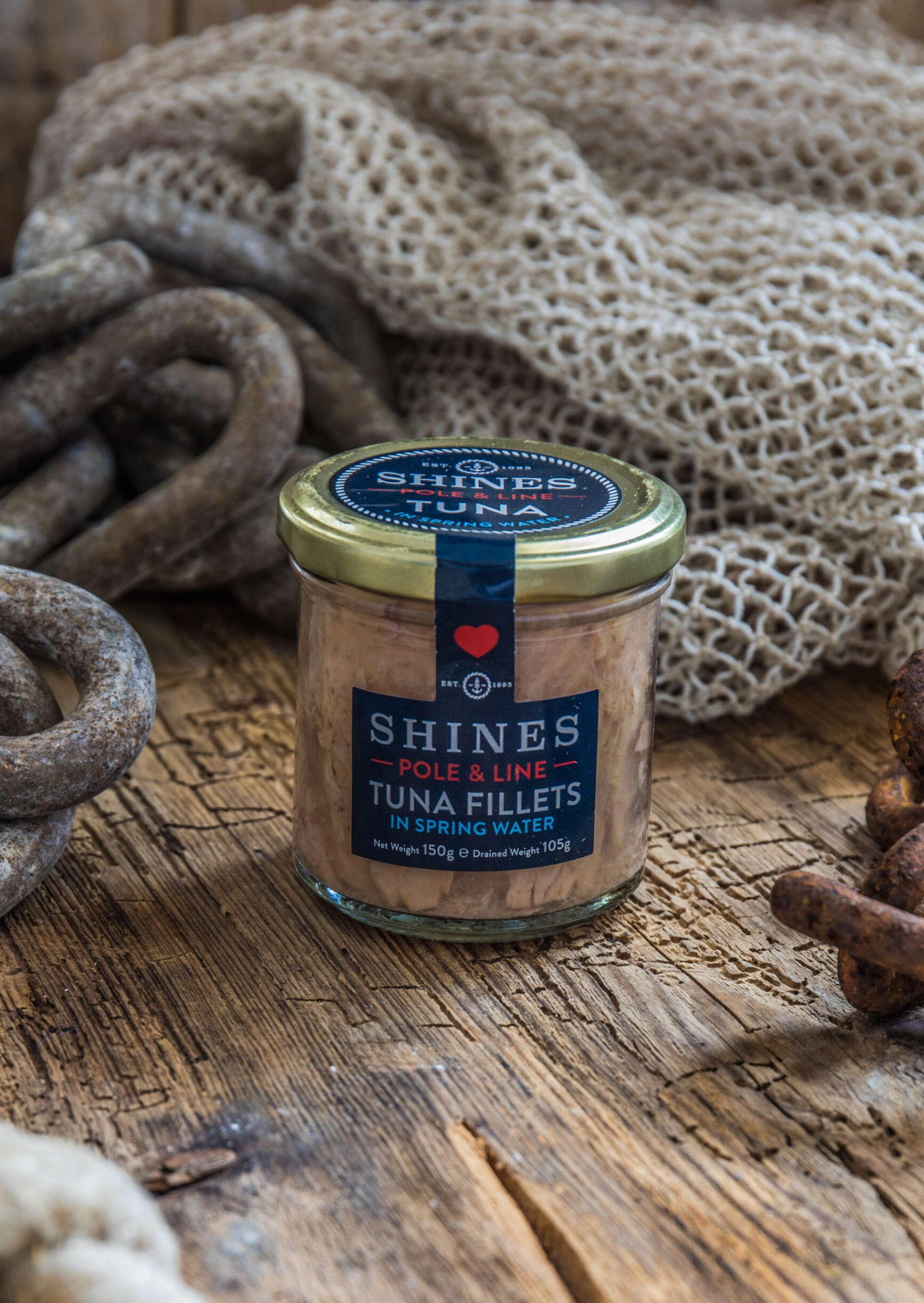 A jar of Shines pole & line tuna fillets in spring water sits on a rustic wooden surface surrounded by fishing nets and rusty chains.