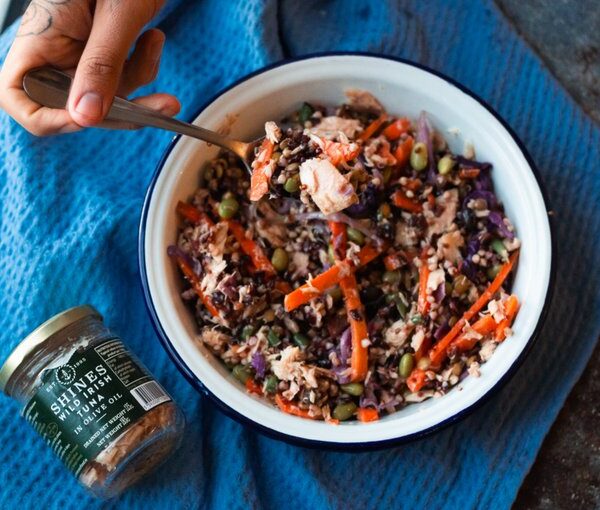 A hand holding a bowl filled with a colourful salad containing shredded vegetables, grains, and tuna. A jar labelled "Shines Wild Irish Tuna in Olive Oil" sits beside the bowl on a blue cloth.