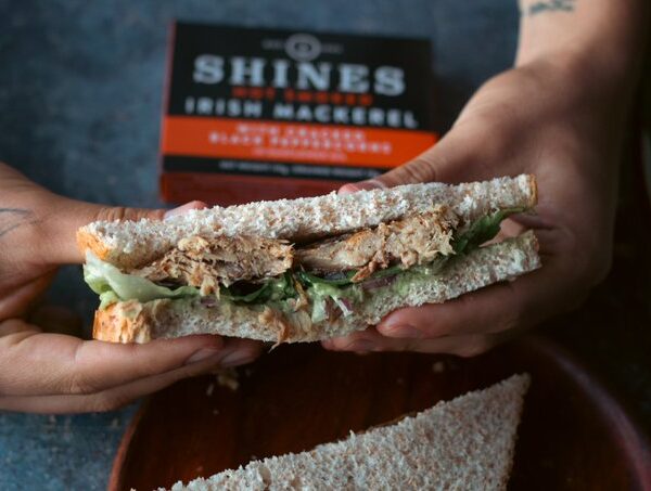 A person with tattooed hands holds a sandwich filled with lettuce and shredded fish on wholemeal bread. A box labelled "Shines Irish Mackerel" is visible in the background.