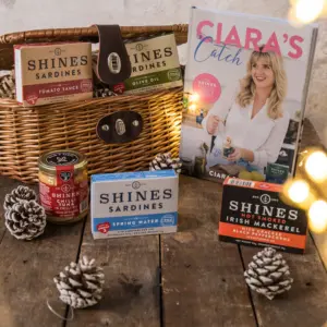 The Cookbook Hamper features Shines sardines, mackerel, salmon tins, pinecones, and Ciara’s Catch cookbook in a wicker basket on rustic wood, with warm fairy lights glowing in the background.