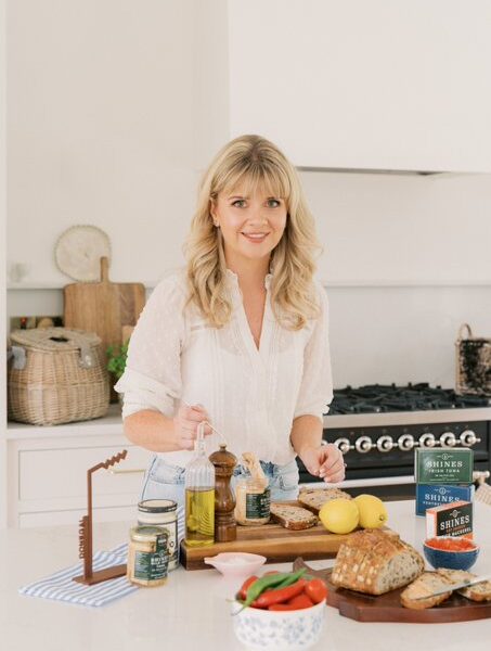 A woman with blonde hair stands in a bright kitchen, preparing food. On the worktop are bread, jars, olive oil, lemons, and a bowl of vegetables. She is smiling and holding utensils over a chopping board.